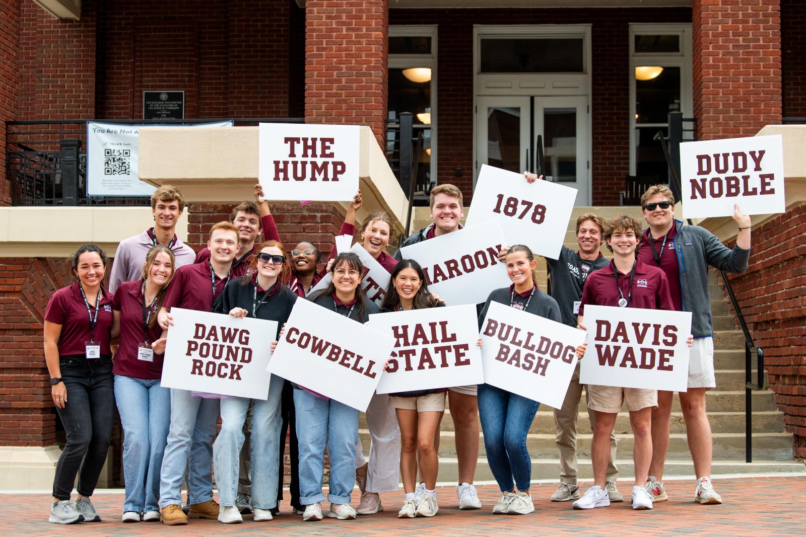 MSU students standing in front of the YMCA building with signs depicting MSU traditions like the Hump, Davis Wade, Cowbell.