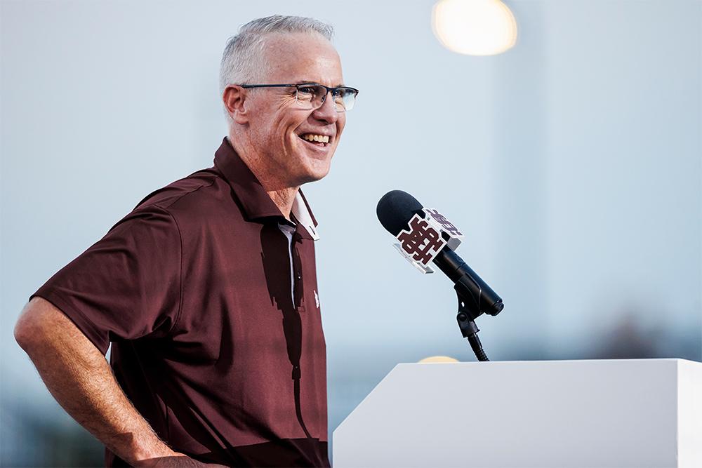 MSU Head Baseball Coach Brian O’Connor smiles while standing at a podium and microphone