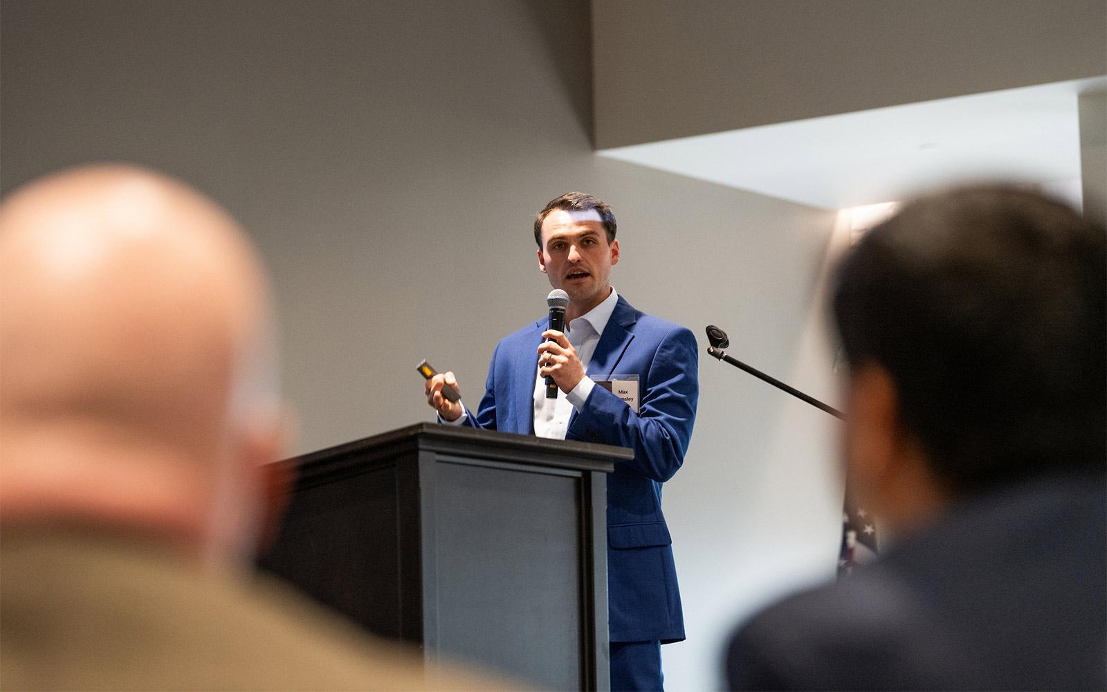 A man holds a microphone and speaks from a podium to listeners in an audience